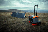 Portable Recon 2000 Power Station with telescoping handle and wheels on dry grass, connected to solar panels under a cloudy sky in a mountainous landscape.