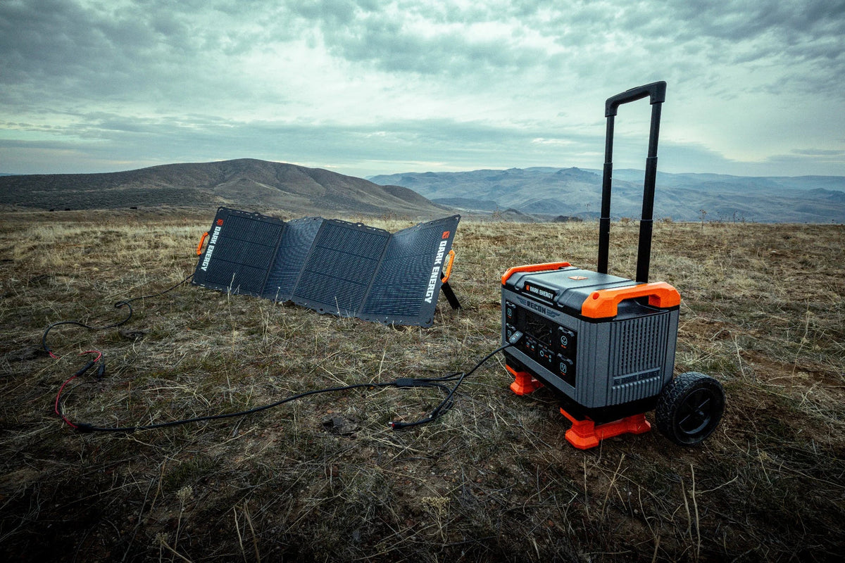 Portable Recon 2000 Power Station with telescoping handle and wheels on dry grass, connected to solar panels under a cloudy sky in a mountainous landscape.