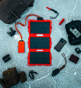 Red-framed tri-fold solar panel connected to an orange power bank, surrounded by camping gear including a black backpack, knife, phone, and pouches on a cracked stone floor.