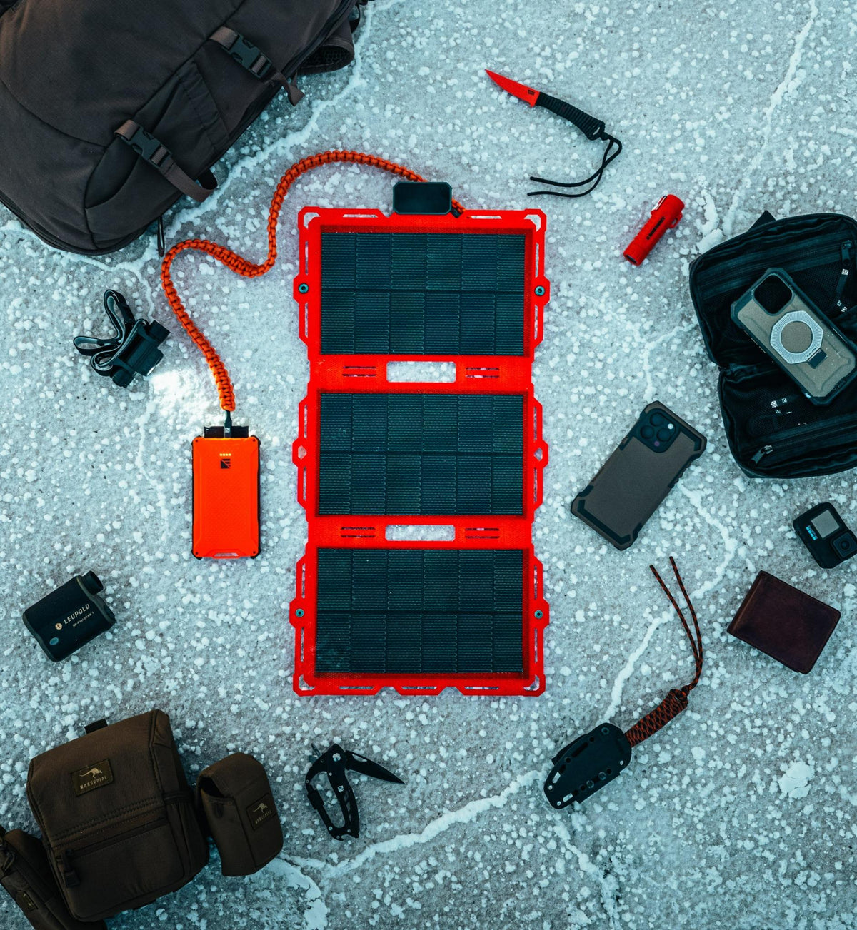 Red-framed tri-fold solar panel connected to an orange power bank, surrounded by camping gear including a black backpack, knife, phone, and pouches on a cracked stone floor.