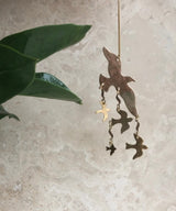 Metallic bird-shaped ornament in warm gold tones with smaller birds hanging below, suspended against a textured beige background near green leaves.