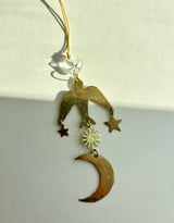 Hammered brass bird ornament hanging by a gold cord with a vintage glass crystal, holding star and sun charms above a crescent moon pendant.