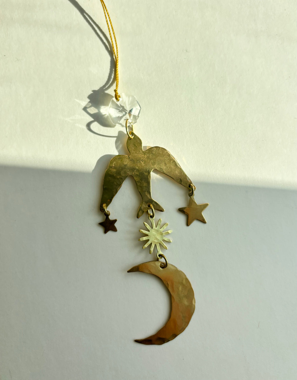 Hammered brass bird ornament hanging by a gold cord with a vintage glass crystal, holding star and sun charms above a crescent moon pendant.