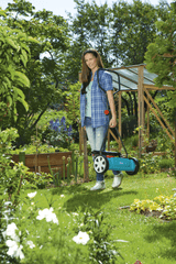 Woman in blue plaid shirt carrying a turquoise and black cylinder lawnmower in a sunny garden with green plants and wooden garden structures.
