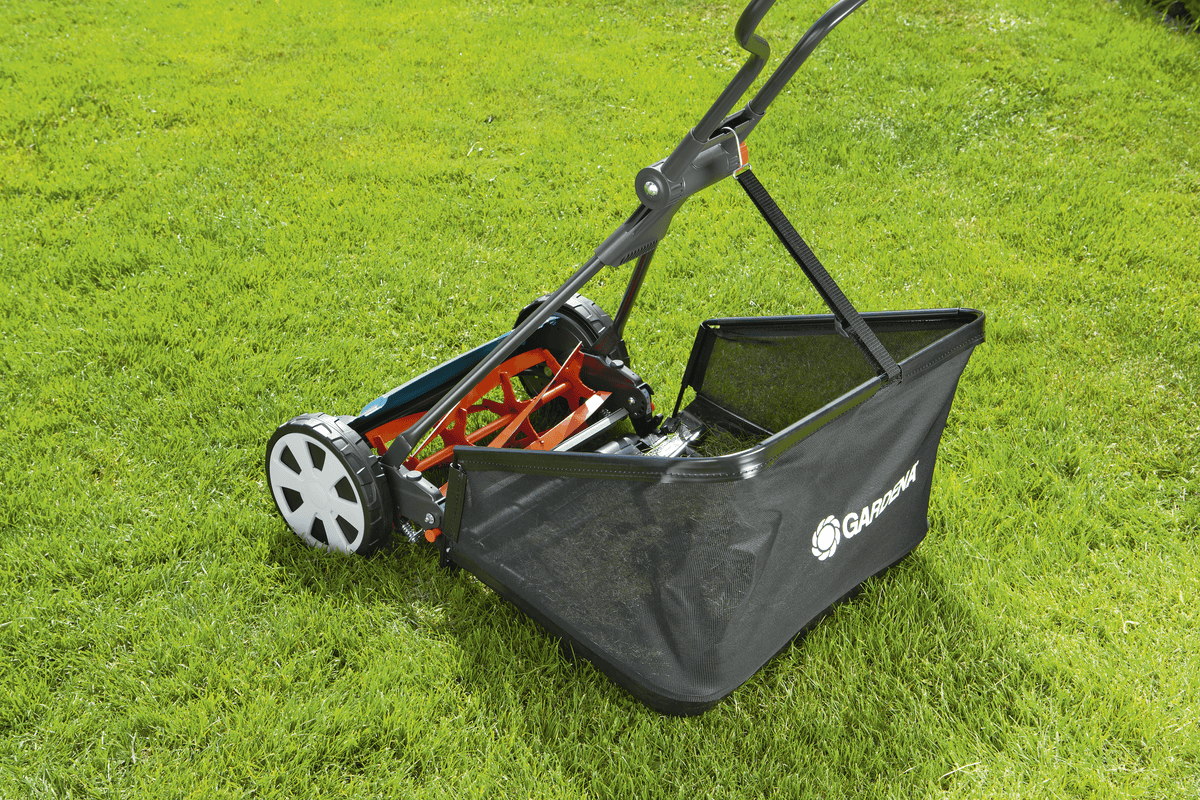 Black manual reel lawn mower with white wheels and black mesh grass catcher, resting on freshly cut green lawn grass under sunlight.