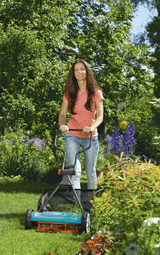 Woman with long brown hair wearing an orange shirt and blue jeans pushing a teal manual grass catcher mower on a green lawn with trees and flowering plants in the background.