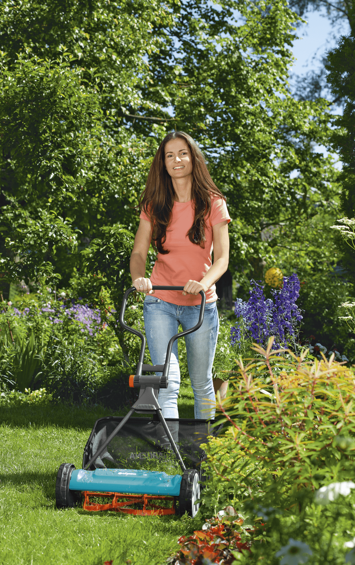 Woman with long brown hair wearing an orange shirt and blue jeans pushing a teal manual grass catcher mower on a green lawn with trees and flowering plants in the background.