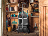 Black lawn leaf collector with blue handles and a mesh bag, positioned in a wooden shed surrounded by gardening tools and boots on shelves.