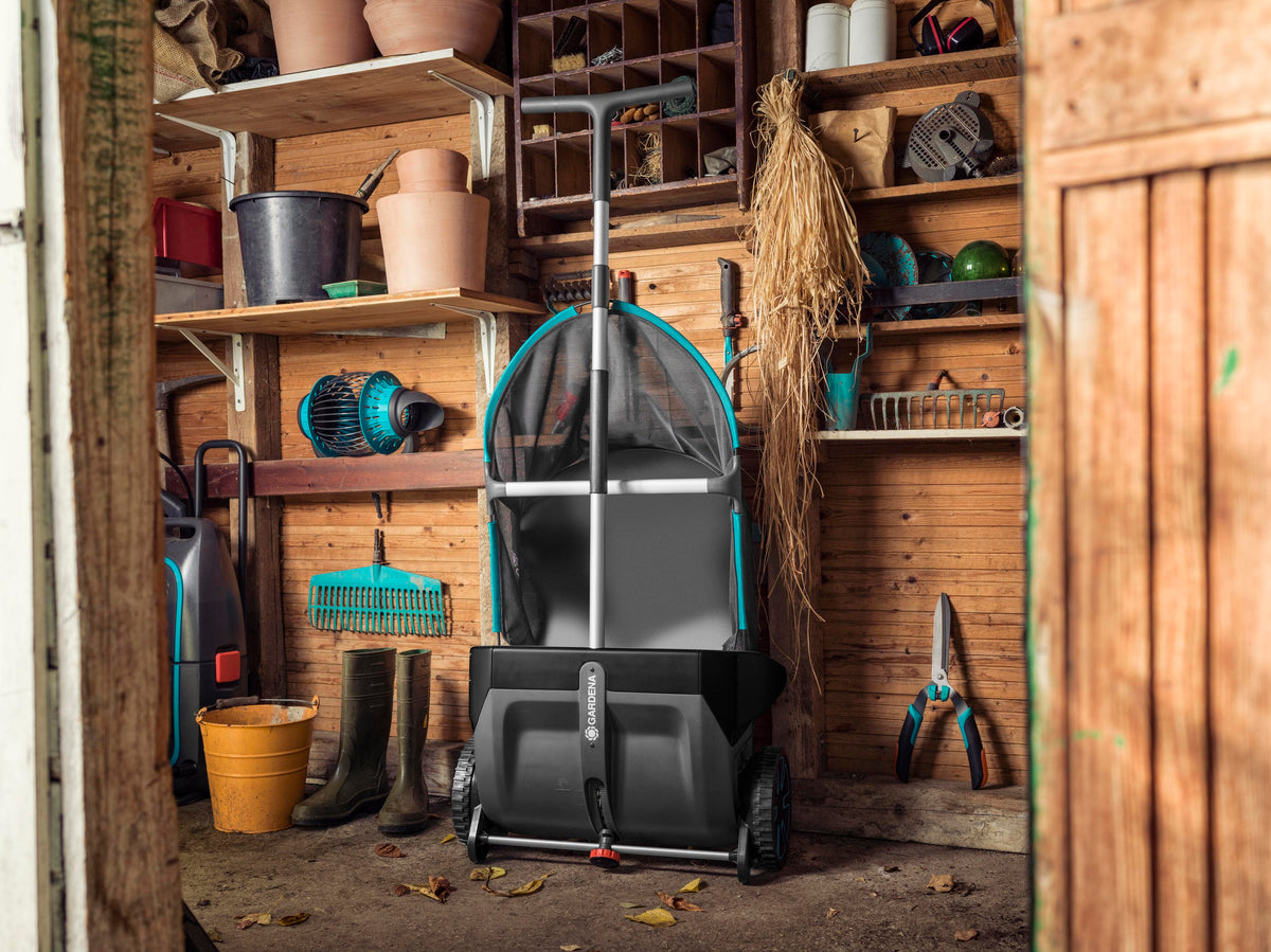 Black lawn leaf collector with blue handles and a mesh bag, positioned in a wooden shed surrounded by gardening tools and boots on shelves.