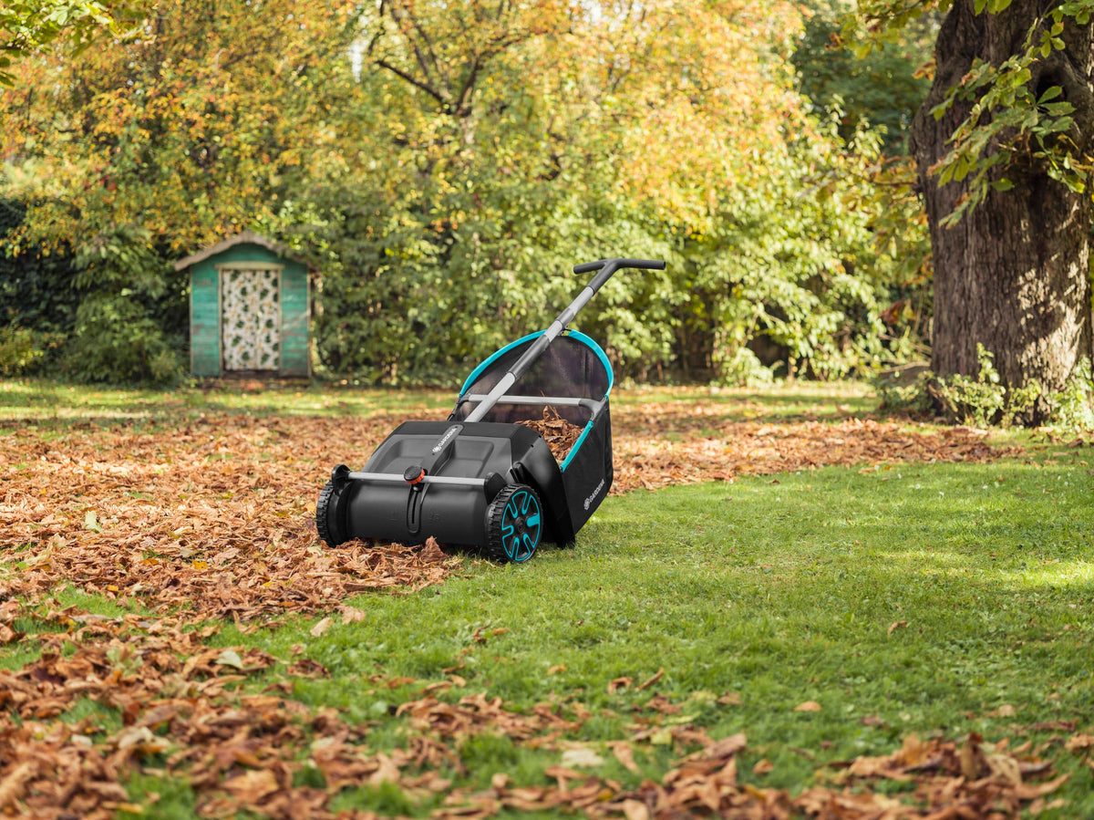 Black and blue lawn leaf collector with handle positioned on grass, with a section of leaves collected, next to a wooded area and small green shed in background.