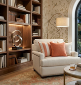 Cream upholstered loveseat with melon-colored striped and solid pillows, set against a wooden built-in bookshelf and decorative lamp near an arched window.