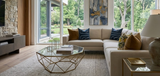 Living room with beige sectional sofa featuring navy and patterned pillows, geometric glass coffee table, light rug, and large window showing green trees outside