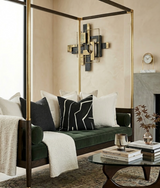 Green velvet sofa with beige and black geometric pillows, draped with a cream throw, positioned under a brass four-poster frame in a neutral-toned living room.