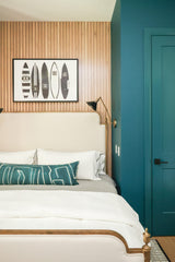 Teal pillow with abstract white lines resting on a beige bed against a wood-paneled wall with black and white surfboard artwork, next to a teal door.