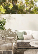 Outdoor seating area with a woven rattan sofa and chair in light beige, decorated with a sage green checkered pillow and a smaller beige pillow with a pink geometric pattern, set against a white wall and shaded by trees.