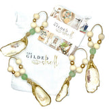 Three oyster shell ornaments edged in gold, each strung on twine with natural wooden beads and a single frosted green bead, arranged on a white drawstring bag and card.