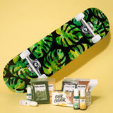 Skateboard with green and yellow leaf pattern deck leaning on a yellow surface, surrounded by Color Creator pigment powders and liquid drops in front.