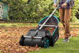 Person pushing a black and turquoise leaf collector with large wheels over a lawn covered in fallen leaves, with green bushes and a shed in the background.