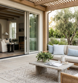 Outdoor seating area with beige cushioned sofa adorned by sky blue woven pillows, a stone coffee table with a plant and open book, set on a neutral patterned rug.