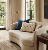 Curved cream sofa with textured beige, patterned cream, and deep blue pillows, beside a wooden side table with stacked books and a brass floor lamp, in a sunlit room with large windows and beige curtains.