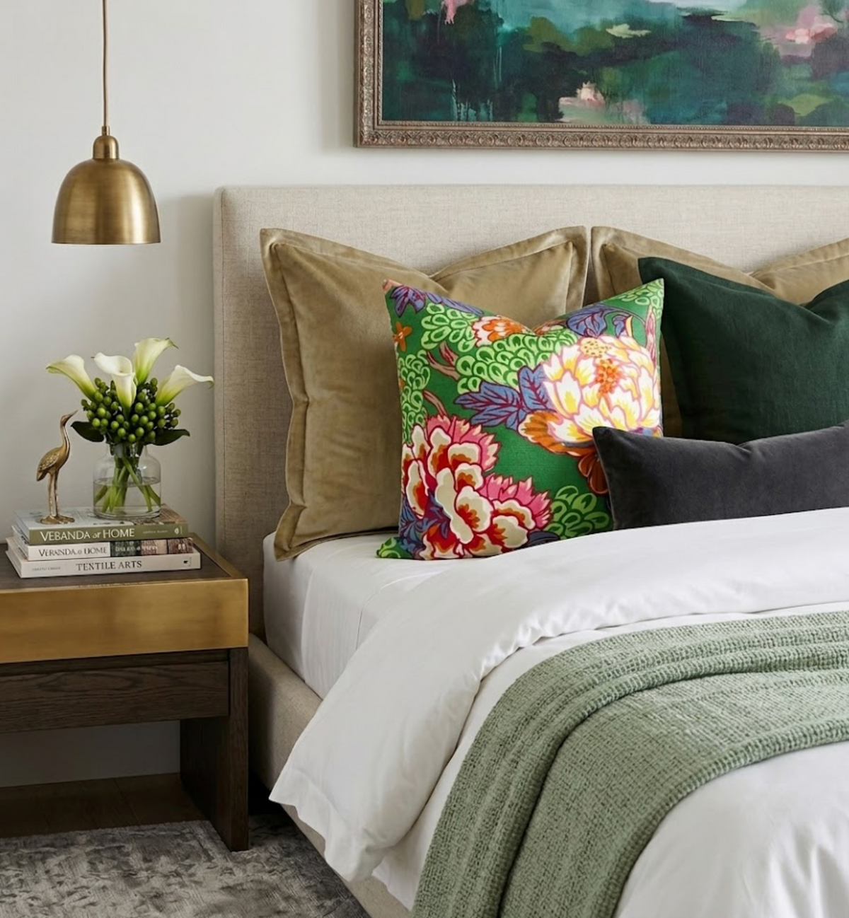 Bed with beige upholstered headboard, white bedding, green knit throw, and vibrant floral decorative pillow among solid-colored pillows, beside wooden nightstand with brass lamp and flowers.