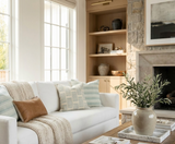 Bright living room with white sofa draped in beige knit throw and patterned pillows, beige built-in shelves, and a stone fireplace with greenery in a ceramic vase on the coffee table.