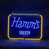 Neon sign featuring blue cursive 'Hamm's' and block 'BEER' text, framed by a glowing yellow rectangular outline and a black plaque with white text above.