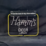 Neon sign with white cursive text 'Hamm's' and block letters 'BEER' inside a yellow square frame, topped by a black plaque reading 'From the land of sky blue waters'.