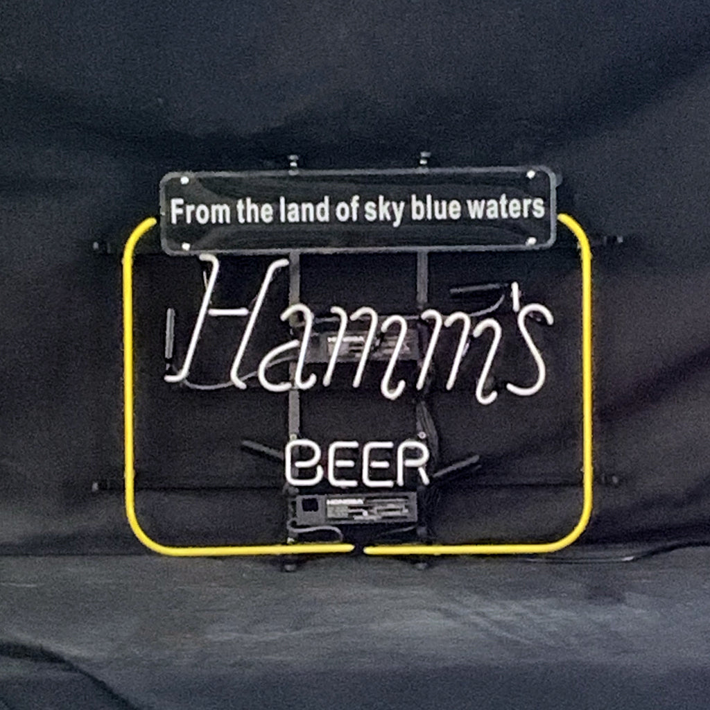 Neon sign with white cursive text 'Hamm's' and block letters 'BEER' inside a yellow square frame, topped by a black plaque reading 'From the land of sky blue waters'.