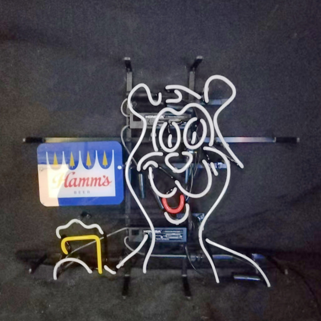 Neon light sign shaped like a cartoon bear holding a beer mug, outlined in white and yellow with a red mouth, featuring a blue Hamm's Beer logo on the left.