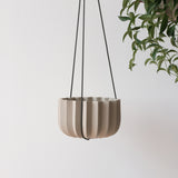 Matte beige ceramic planter with vertical ridges suspended by thin black cords against a plain white background with a green leafy plant on the right.