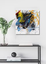 Square abstract canvas with bold blue, black, yellow, and orange paint strokes and splatters on a white background, mounted on a white wall above a minimalist black metal table.