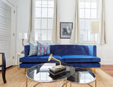 Bright living room with a deep blue velvet sofa adorned by two patterned pillows, a round glass coffee table featuring a vintage brass gramophone, and tall windows with neutral curtains.