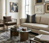 Living room with charcoal ikat patterned pillow on beige sofa, flanked by brown velvet chairs and a glass coffee table with curved bronze base on a patterned rug.