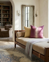Daybed with beige cushion, yellow and pink patterned ikat pillows, and a gray throw, placed in a bright room with a rustic mirror and bookshelf in the background.