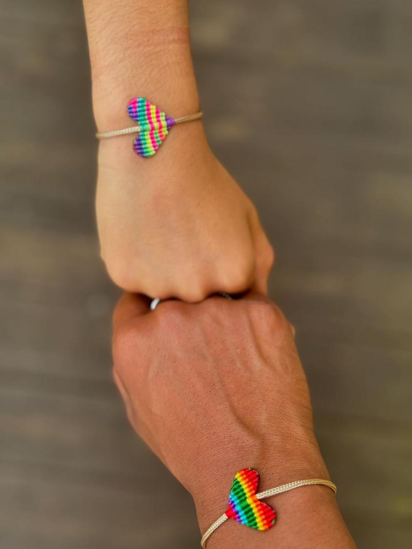 Two hands wearing beige cord bracelets with small rainbow-striped heart charms, joined together against a blurred brown background.
