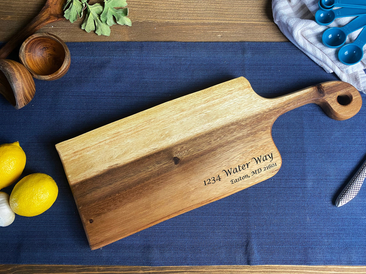 Rectangular wooden bread board with a rounded handle featuring a hole, engraved with an address, lying on a blue cloth with lemons and wooden bowls nearby.
