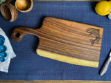 Small live edge wooden cutting board with handle and engraved fish design on one corner, resting on a textured blue cloth with lemon and kitchen items nearby.