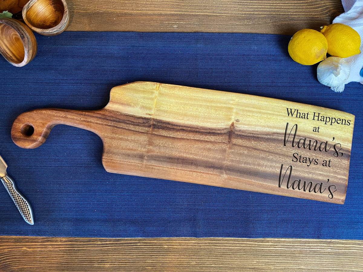 Long wooden handled bread board with natural grain and 'What Happens at Nana's Stays at Nana's' engraved, lying on a blue cloth on a wooden table.