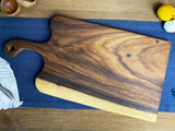 Rectangular live-edge wooden cutting board with rounded handle, dark and light wood grain variations, resting on a blue cloth on a wooden table.