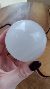 Hand holding a smooth, polished white selenite sphere with subtle translucent and milky swirls, above a wooden surface with natural grain and grooves.