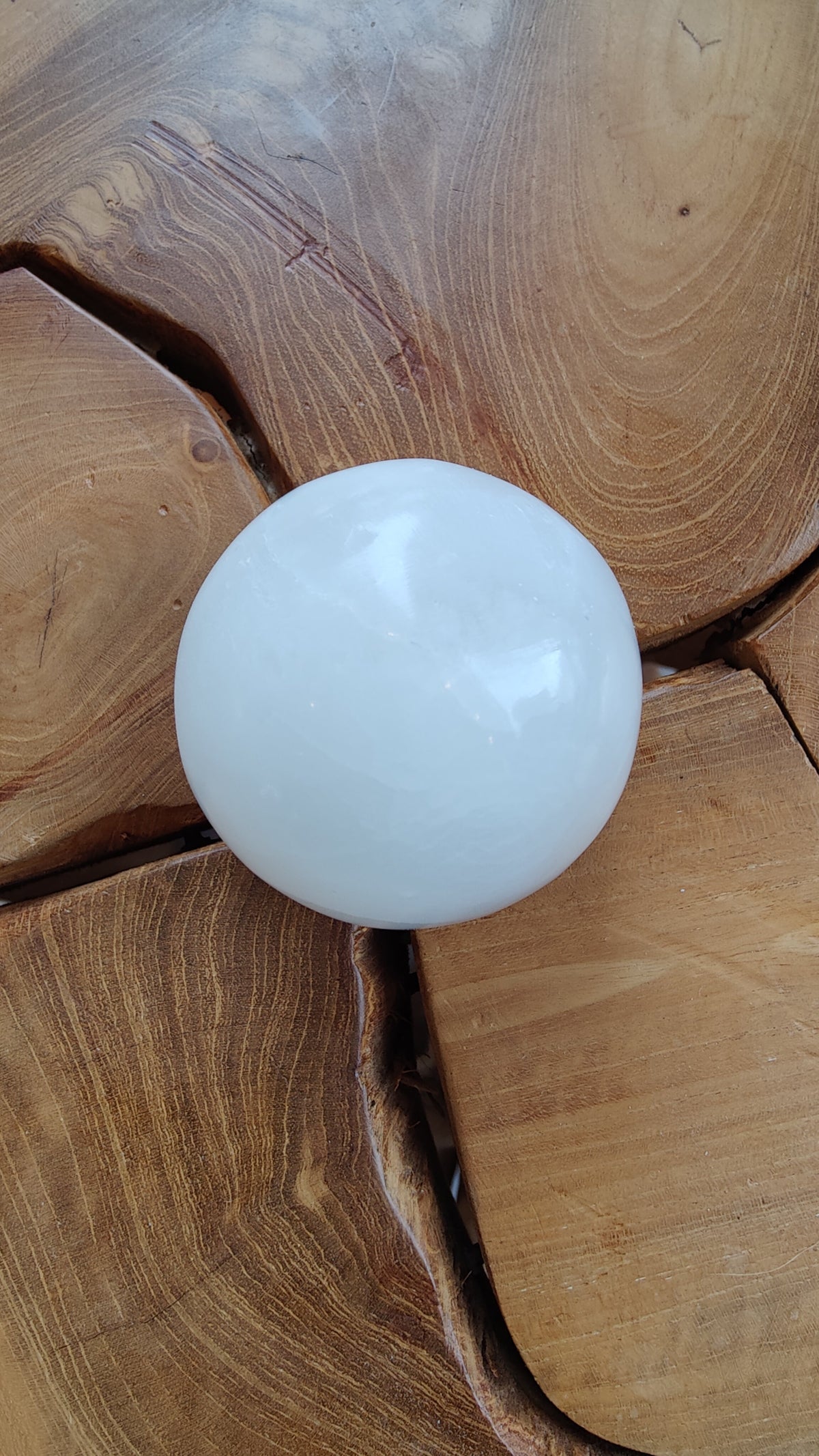 Smooth white selenite sphere measuring approximately 6cm resting on a light brown wooden surface with natural grain and radial patterns.