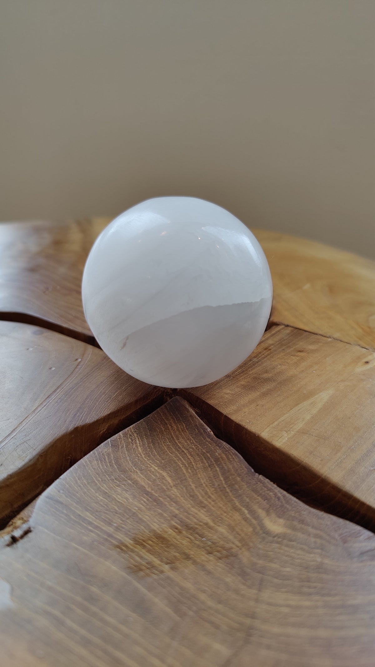 Smooth white selenite sphere with subtle striations, resting on a wooden surface with natural cracks and a warm brown tone, photographed against a neutral background.
