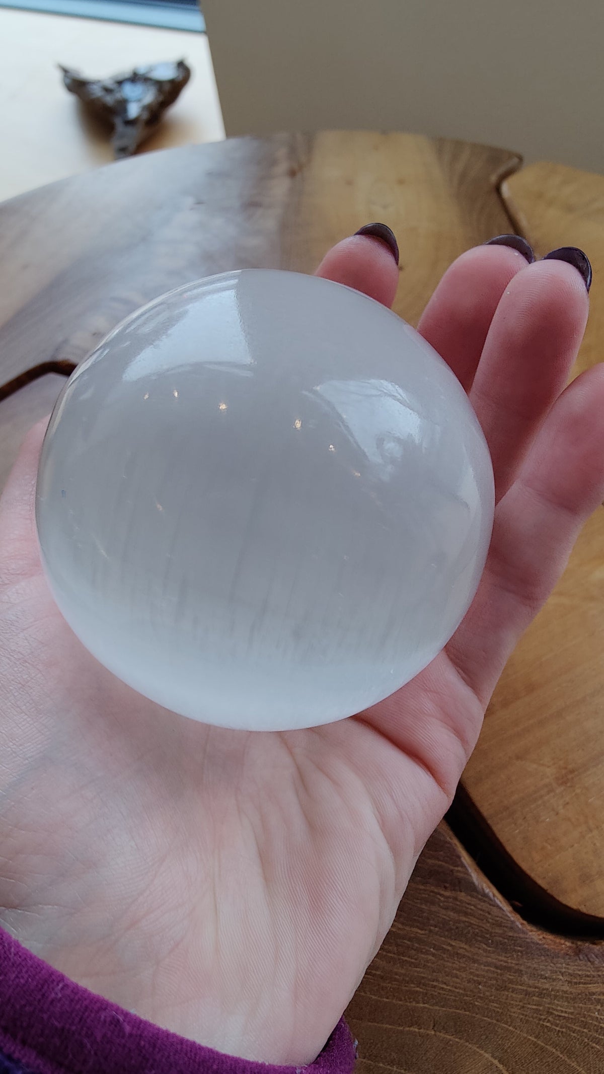 Hand holding a smooth, translucent white selenite sphere with subtle linear striations, resting above a natural wood surface with warm tones and irregular edges.