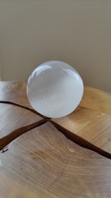 Clear, polished selenite sphere with subtle internal striations resting on a natural wooden surface with deep grain and a prominent split.