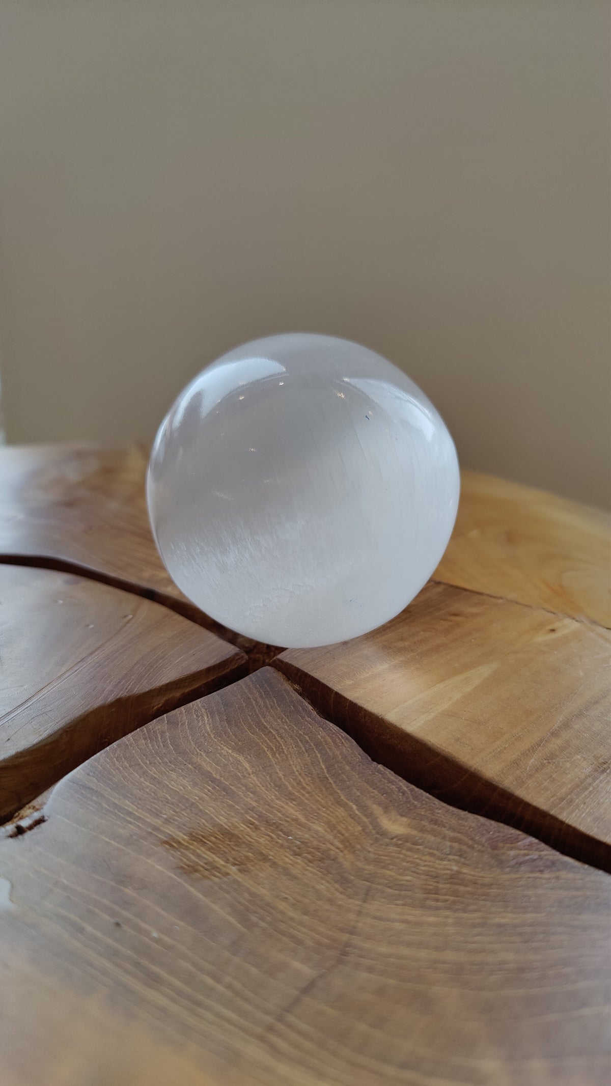 Clear, polished selenite sphere with subtle internal striations resting on a natural wooden surface with deep grain and a prominent split.