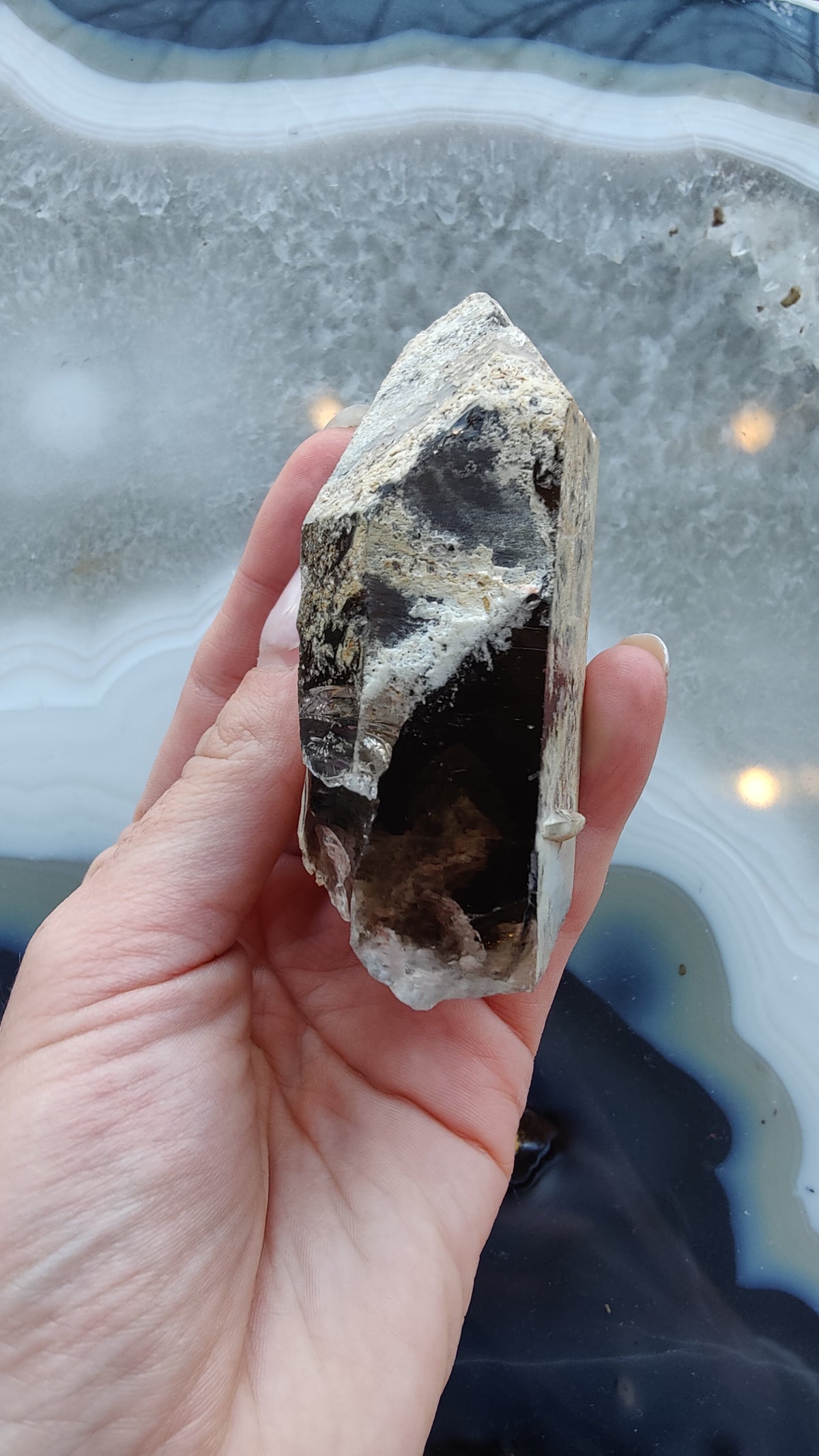 Hand holding a rough smoky quartz crystal with black lodalite inclusions against a gray and blue agate stone background with curved bands.