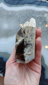 Hand holding a pointed smoky quartz crystal with visible lodolite inclusions, showing natural brown and translucent areas against a blue-gray agate background.
