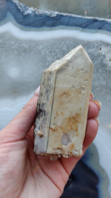 Hand holding a pointed, translucent smoky quartz crystal with visible lodolite inclusions and rough textured base against a blue and white agate background.