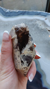 Hand holding a pointed smoky quartz crystal with visible lodalite mineral inclusions and semi-transparent brown and beige tones against a blue and white agate slab background.